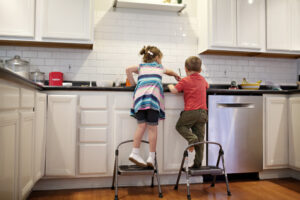 a young boy and girl on stools working at the kitchen sink